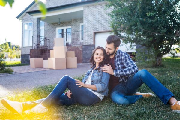 man and woman sitting on the lawn in the front of their house with moving boxes behind them